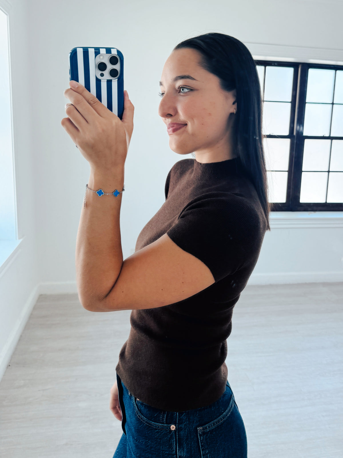 A woman takes a mirror selfie in a bright room, wearing a brown short-sleeve mock neck top and blue jeans. She is holding a phone with a blue and white striped case, and wears a blue beaded bracelet on her wrist.