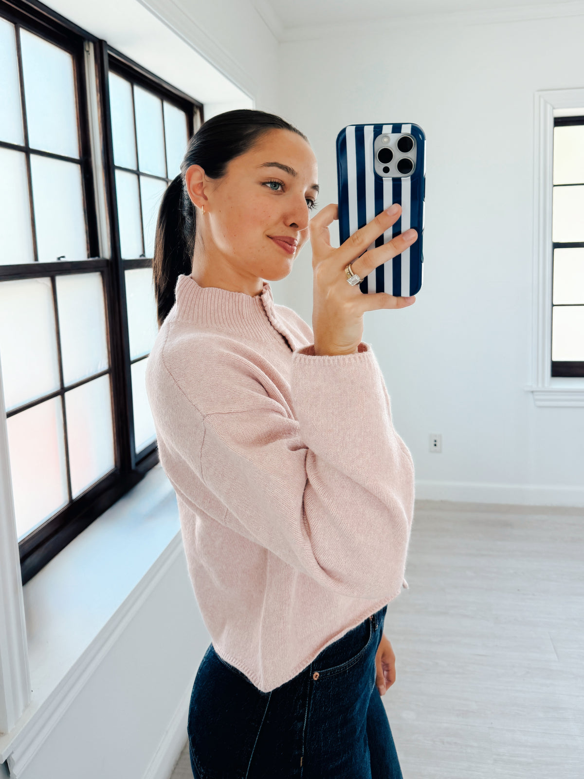 A woman takes a mirror selfie in a bright, modern room with white walls and large black-framed windows. She is wearing a light pink ribbed knit mock-neck sweater and dark blue jeans, holding a blue and white striped phone case.