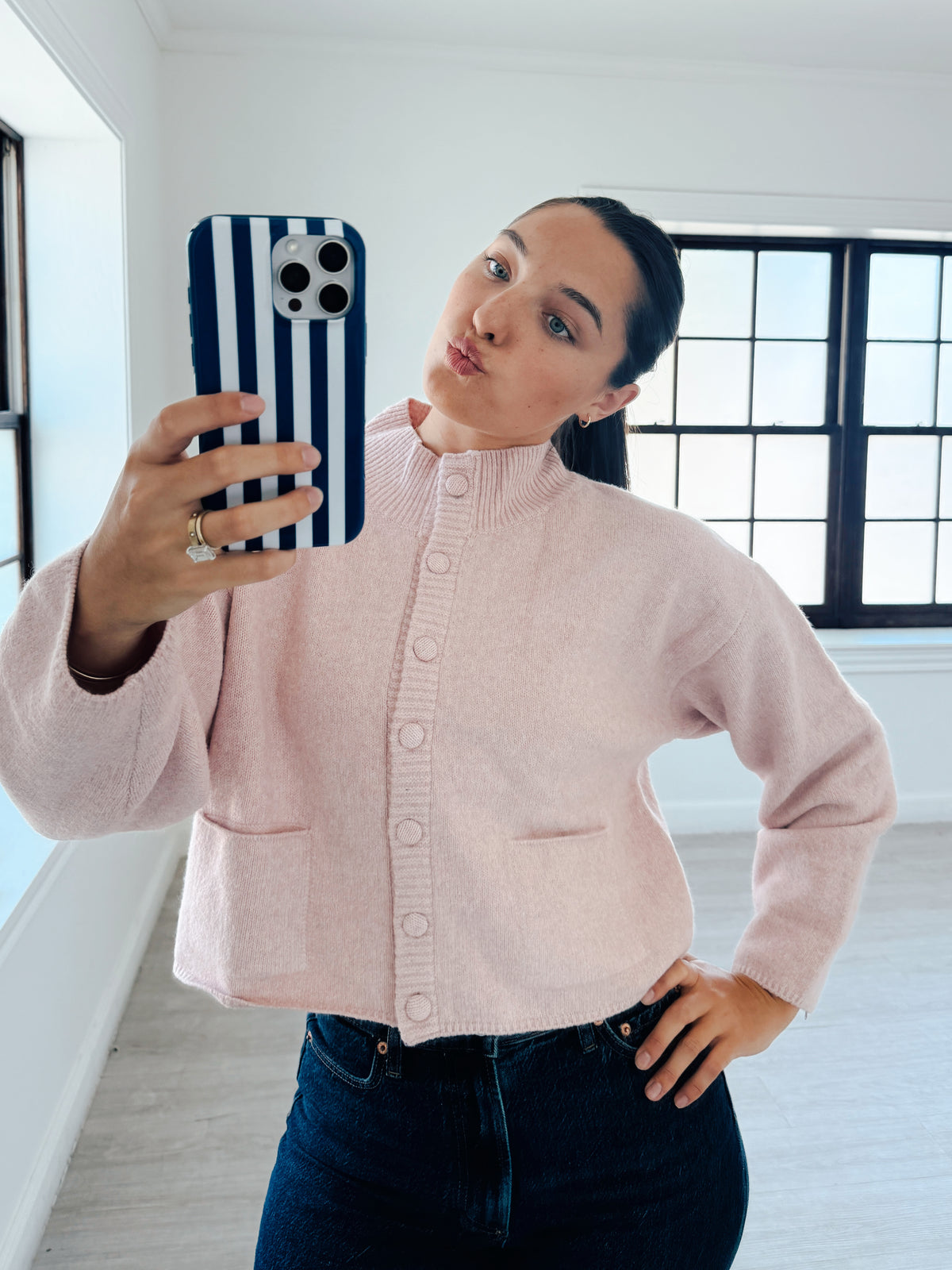 A woman taking a mirror selfie wearing a light pink knit button-up cardigan and dark blue jeans, holding a phone with a navy and white striped case in a bright room with large black-framed windows.