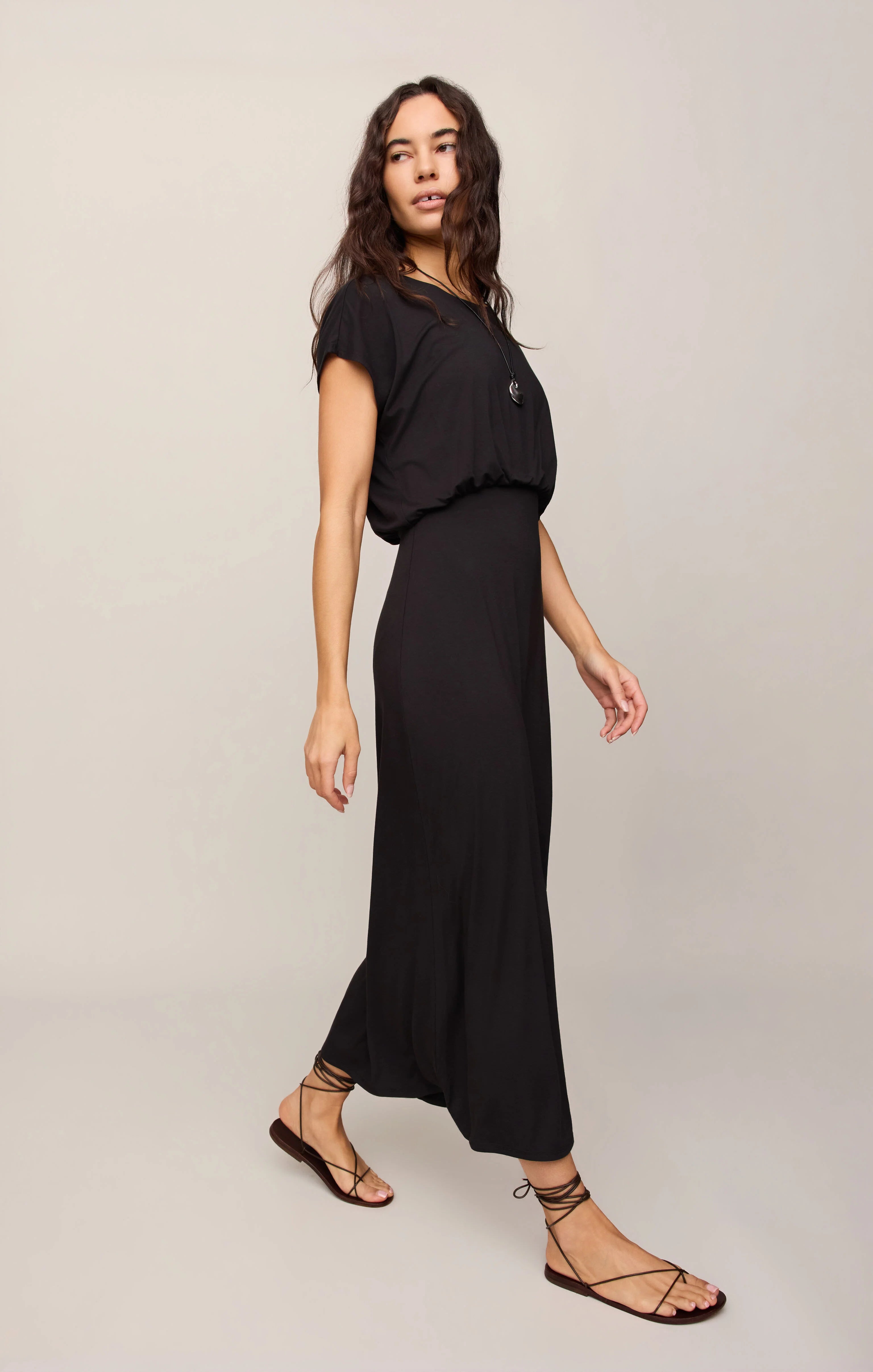 A woman modeling a black short-sleeve maxi dress with a gathered waist and flowing skirt, paired with black strappy flat sandals, against a light gray studio background.