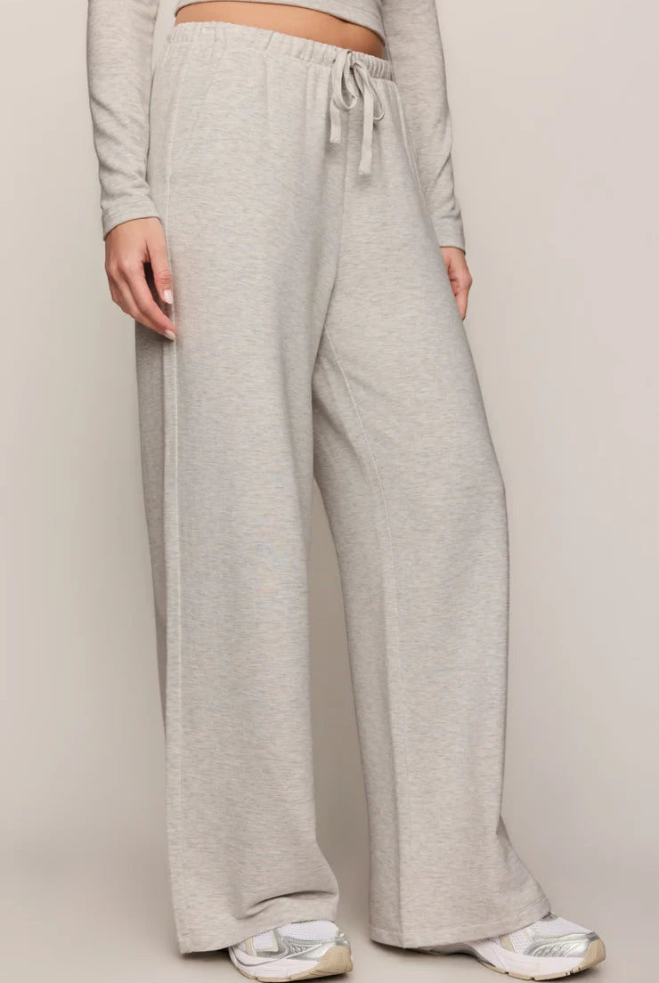 A person wearing light heather gray wide-leg sweatpants with an elastic drawstring waist, paired with a matching long-sleeve crop top and white sneakers, against a plain light beige studio background.