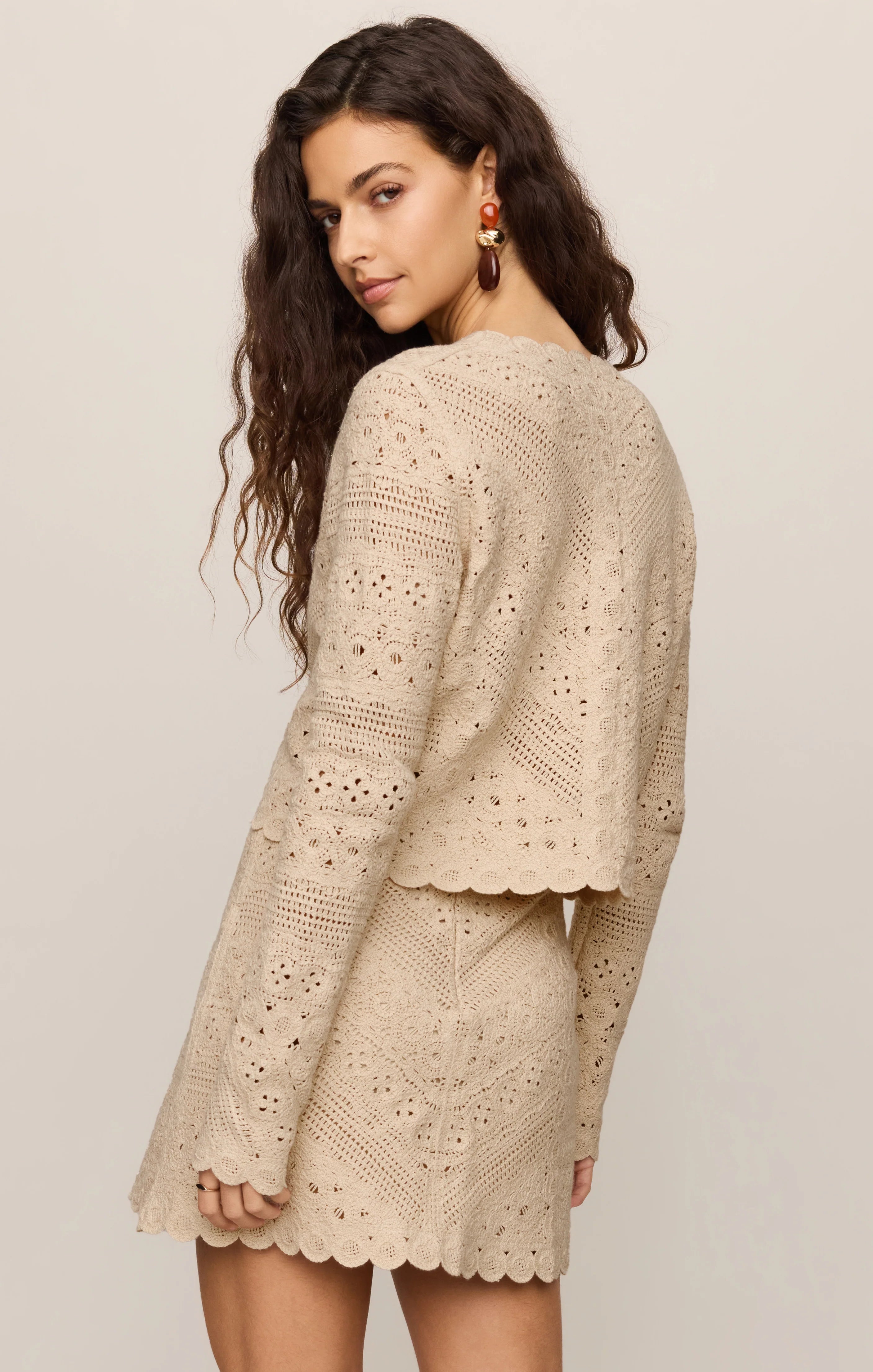 A woman modeling a beige eyelet crochet knit set consisting of a cropped long-sleeve cardigan and a matching mini skirt, shown from a three-quarter back angle against a light beige studio background.
