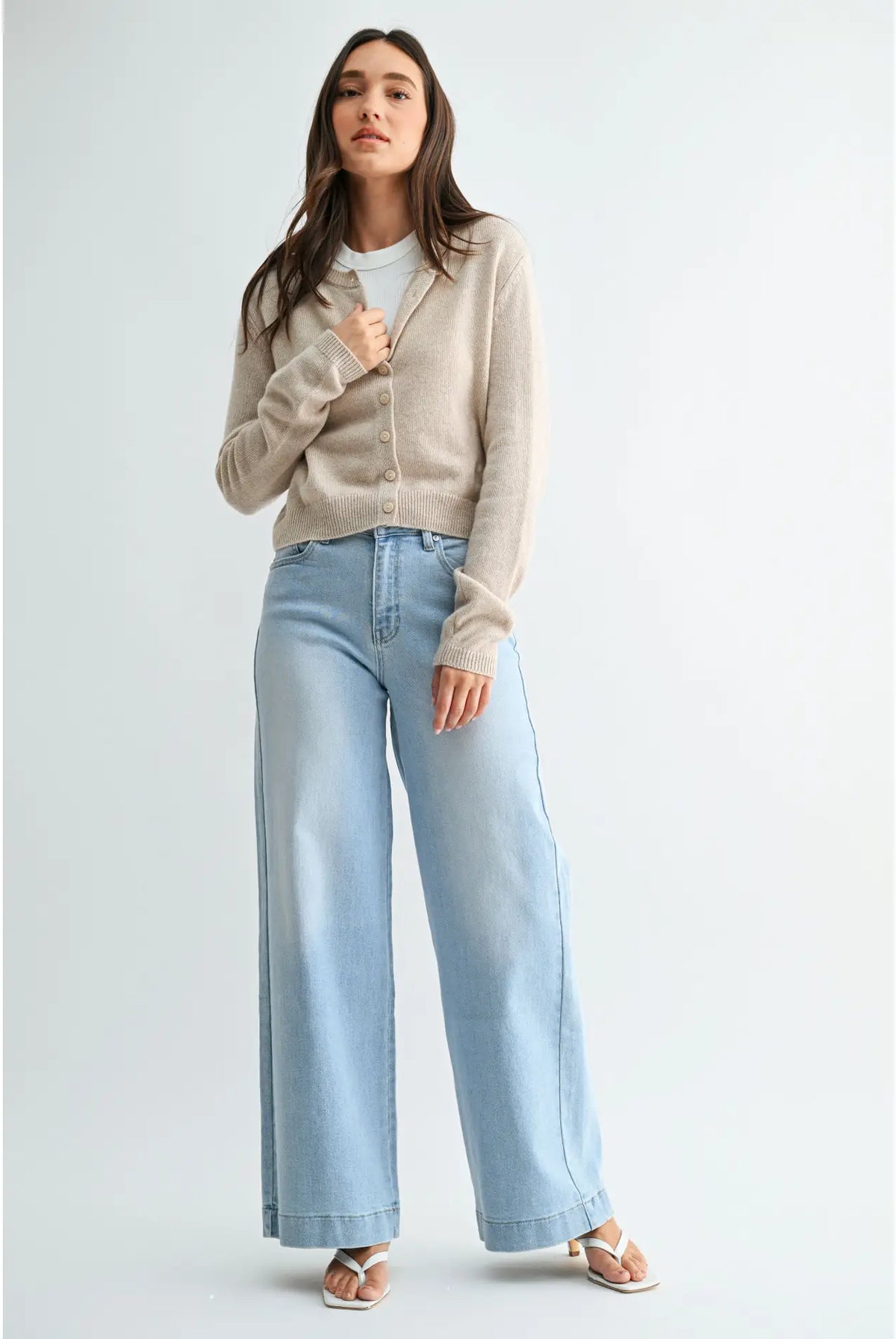 A woman modeling a light beige knit cardigan over a white top, paired with light-wash wide-leg jeans and beige sandals, against a plain light gray studio background.