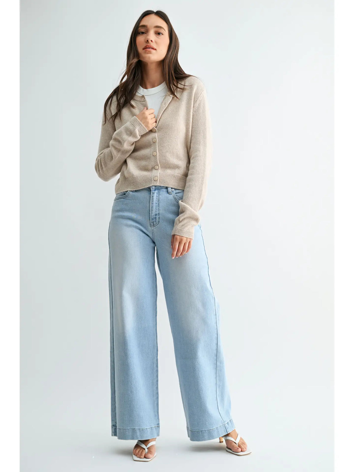 A woman modeling a light beige knit cardigan over a white top, paired with light-wash wide-leg jeans and beige sandals, against a plain light gray studio background.