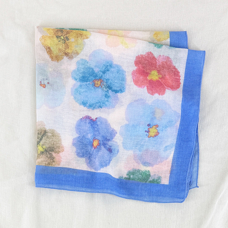 A square white scarf with a colorful watercolor floral print and a solid blue border, folded on a textured white fabric background.