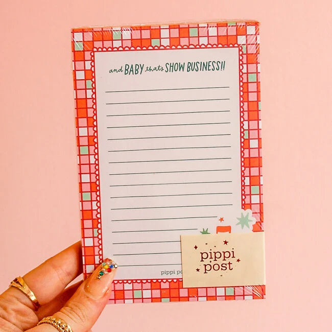 Notepad with a checkered border held by a hand on a pink background, featuring the brand 'pippi post'.