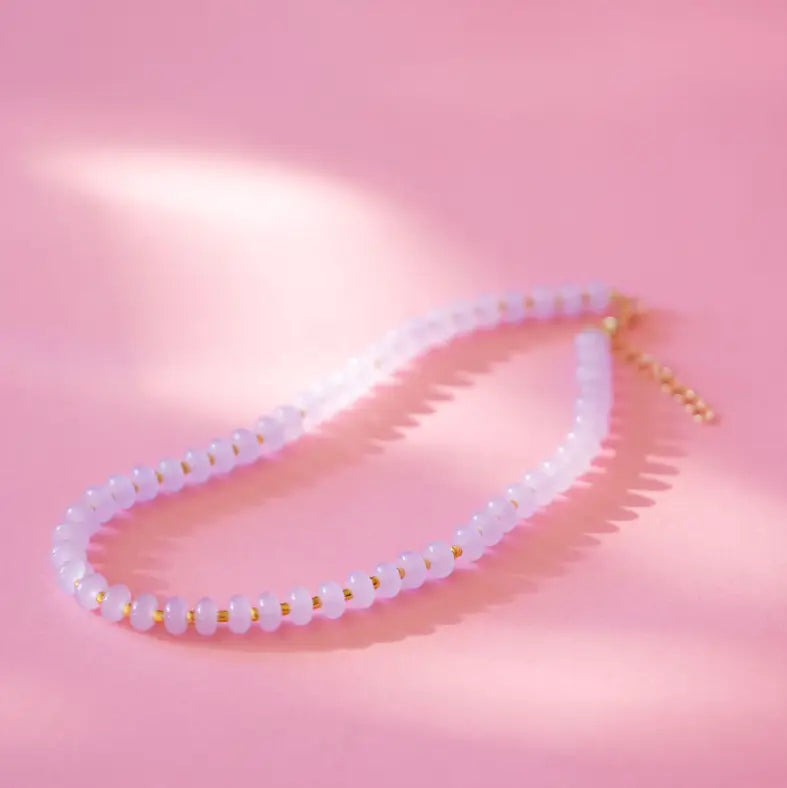 A delicate beaded necklace with pale pink translucent beads separated by small gold spacer rings, displayed on a soft pink background with artistic shadows.
