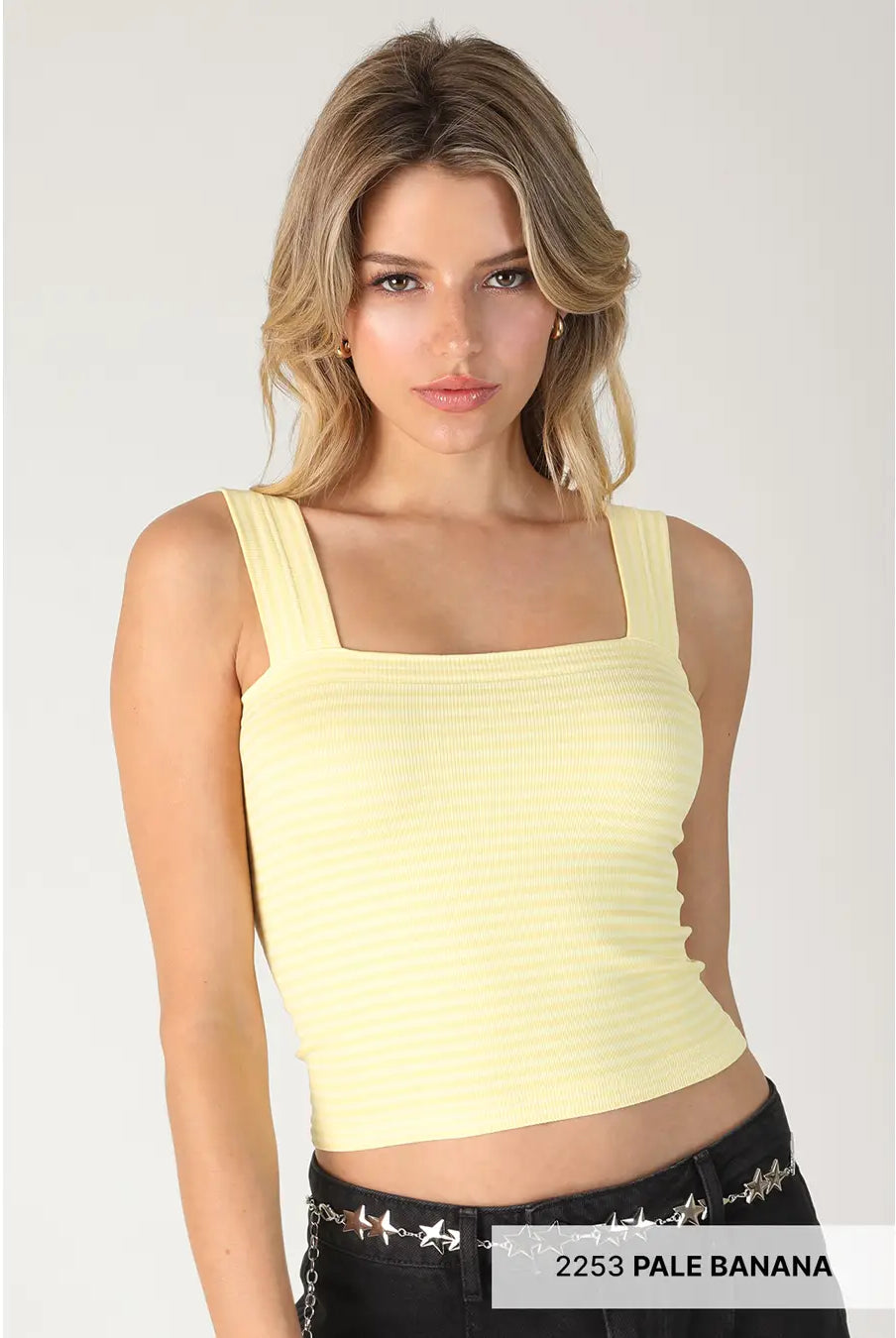 A woman modeling a pale yellow ribbed crop top with a square neckline and wide straps, paired with black studded jeans against a light gray studio background.