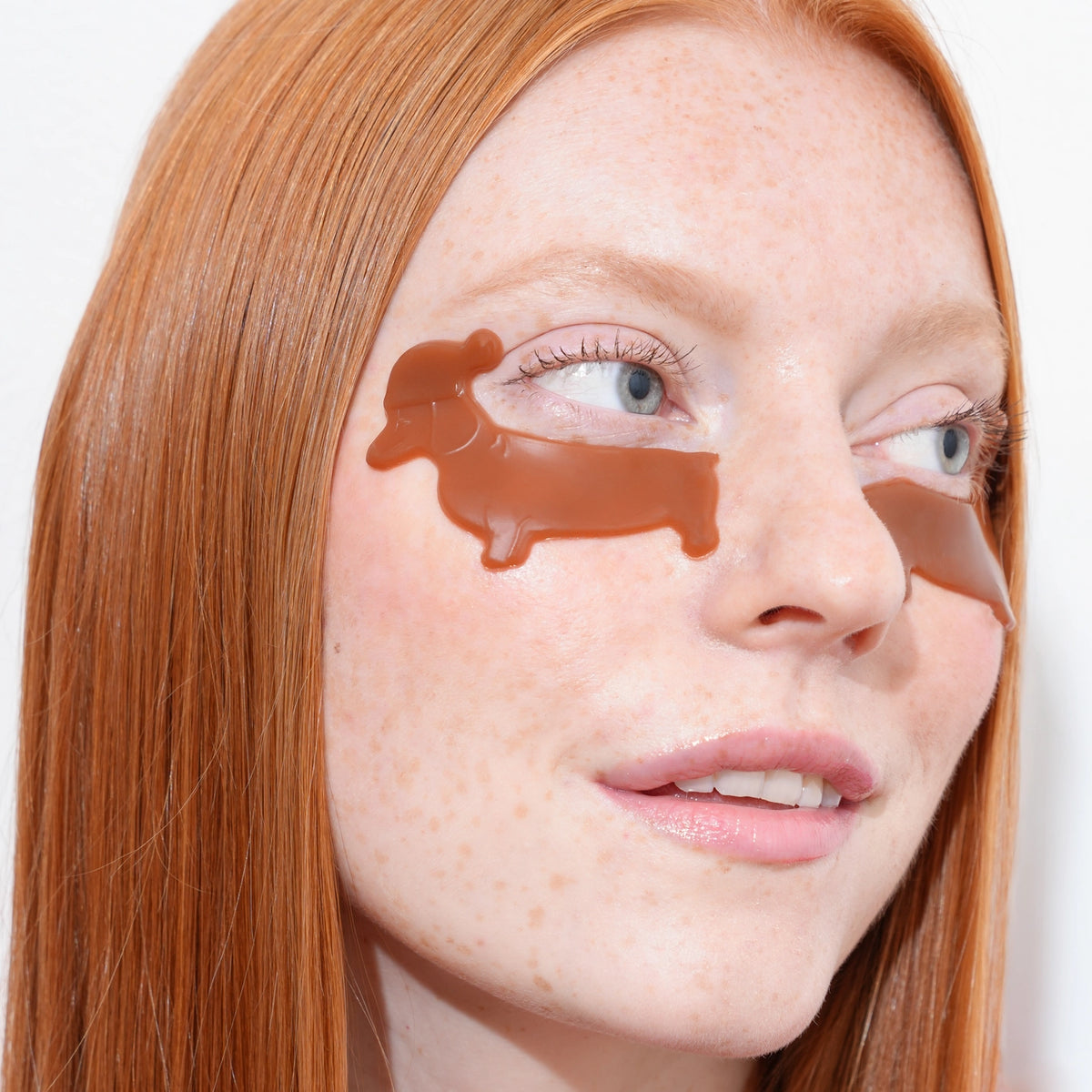 Close-up of a woman with red hair wearing brown silicone eye patch covers under her eyes, shown against a plain light background.