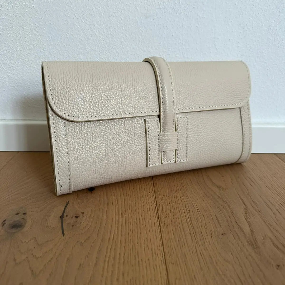 Beige leather clutch bag on a wooden floor with a white wall background