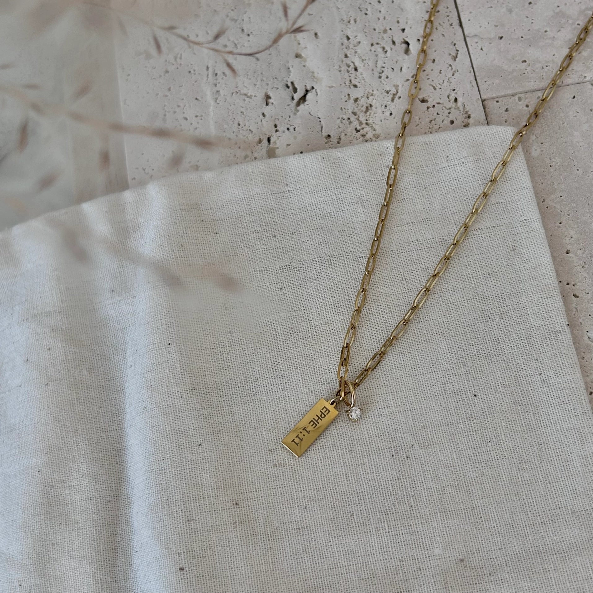 A gold chain necklace with a small rectangular engraved gold tag pendant and a tiny clear stone charm, laid on a textured off-white linen fabric.