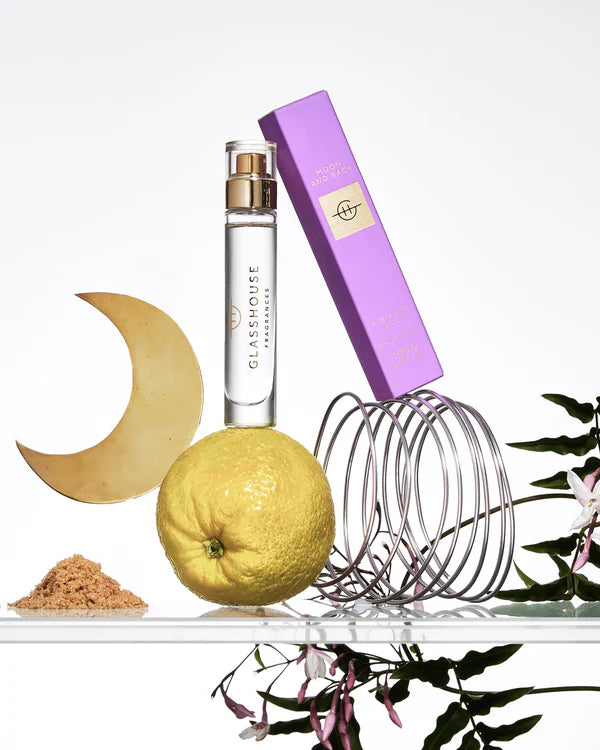 Glasshouse perfume bottle with purple cap, lemon, moon-shaped object, and floral elements on a white background