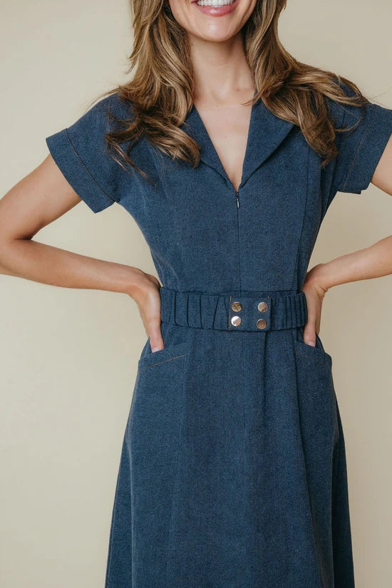 A woman wearing a dark blue denim short-sleeve dress with a buttoned waist and front pockets, posed against a solid beige background.