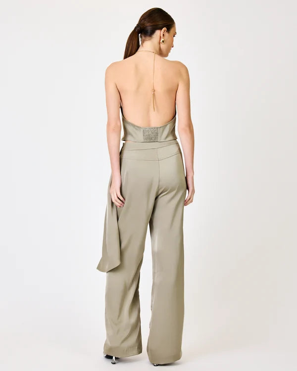 A woman shown from the back wearing a light taupe open-back jumpsuit with wide-leg trousers and a draped side detail, photographed against a plain light gray studio background.