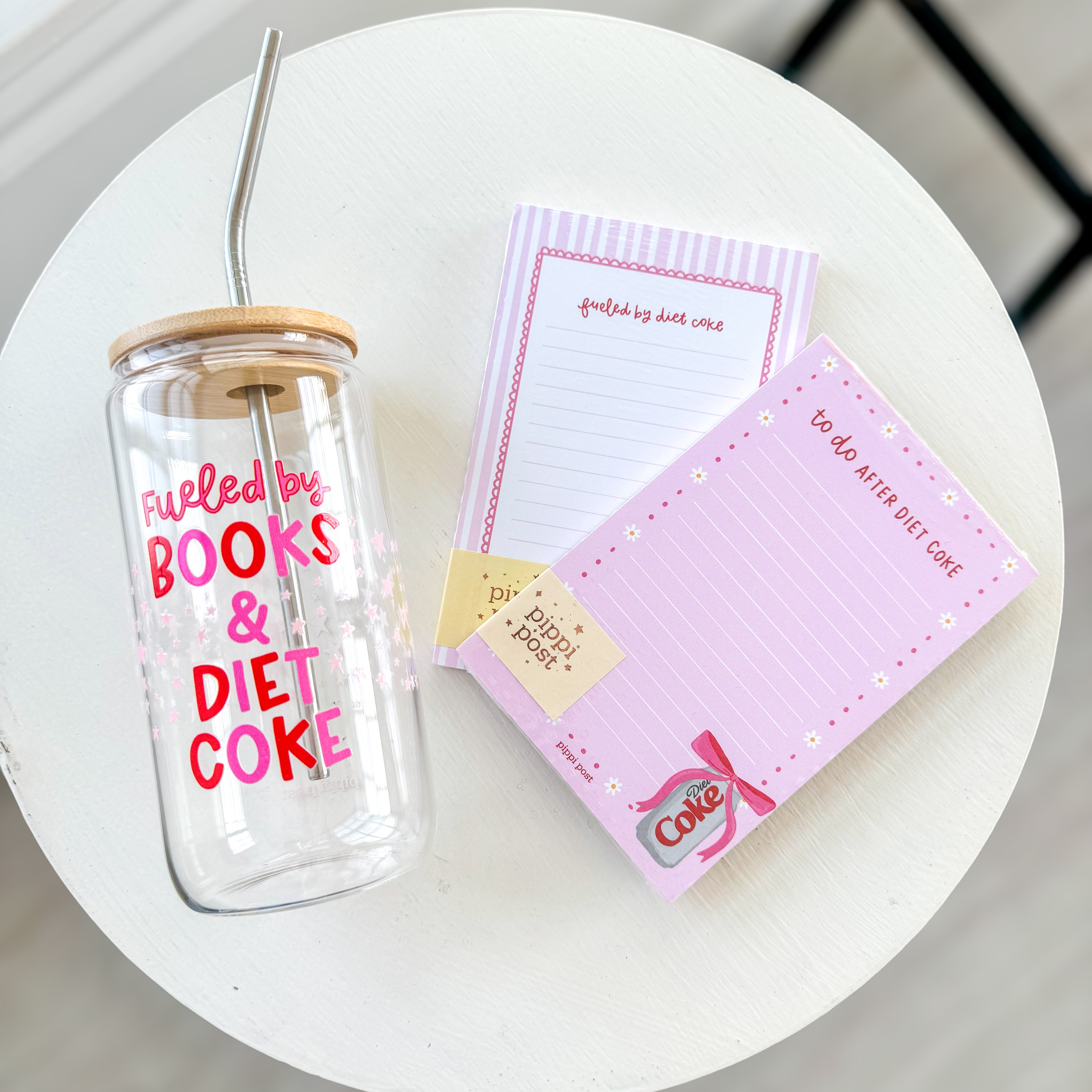 A clear glass tumbler with a bamboo lid and straw reading “Fueled by Books & Diet Coke” sits on a white table beside two pink notepads—one striped and one with a bow and Diet Coke can illustration—styled as part of a girly, aesthetic desk setup.