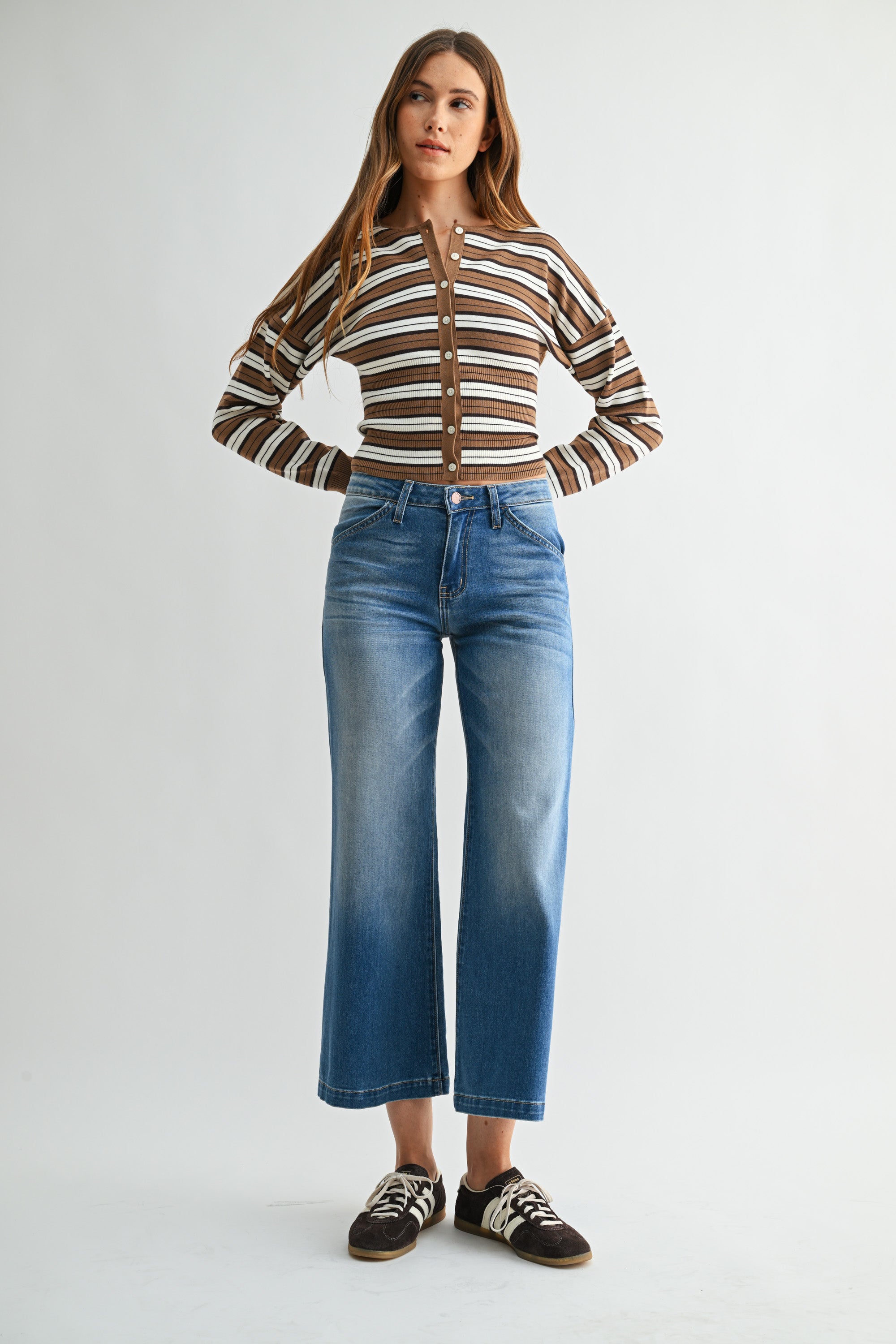 A woman modeling a brown and white striped long-sleeve knit top with a button-up front, paired with blue cropped jeans and brown-and-white sneakers, against a light gray studio background.