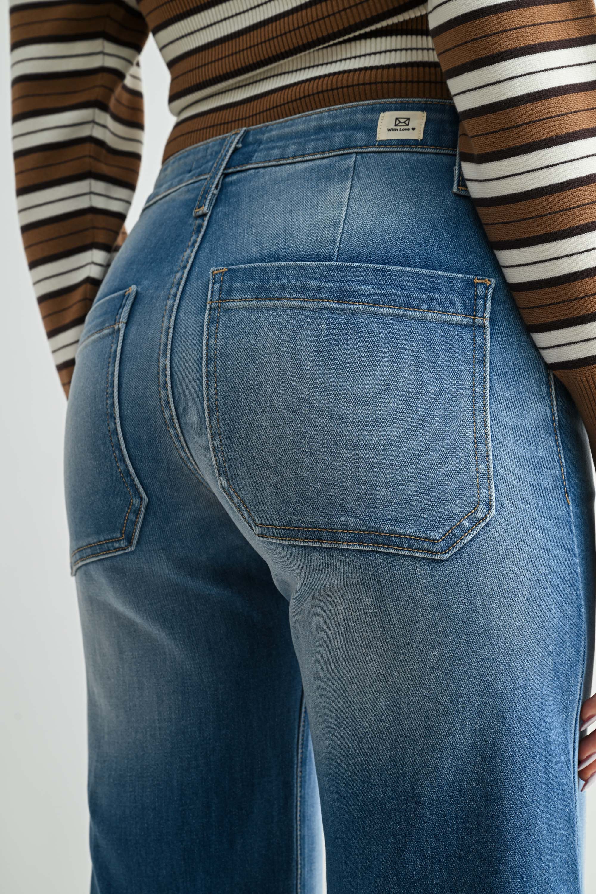 Close-up back view of a person wearing medium-wash blue denim jeans and a brown, white, and black striped long-sleeve top, showing the back pocket and waistband details.