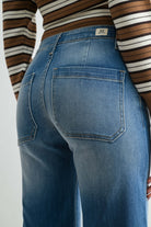 Close-up back view of a person wearing medium-wash blue denim jeans and a brown, white, and black striped long-sleeve top, showing the back pocket and waistband details.