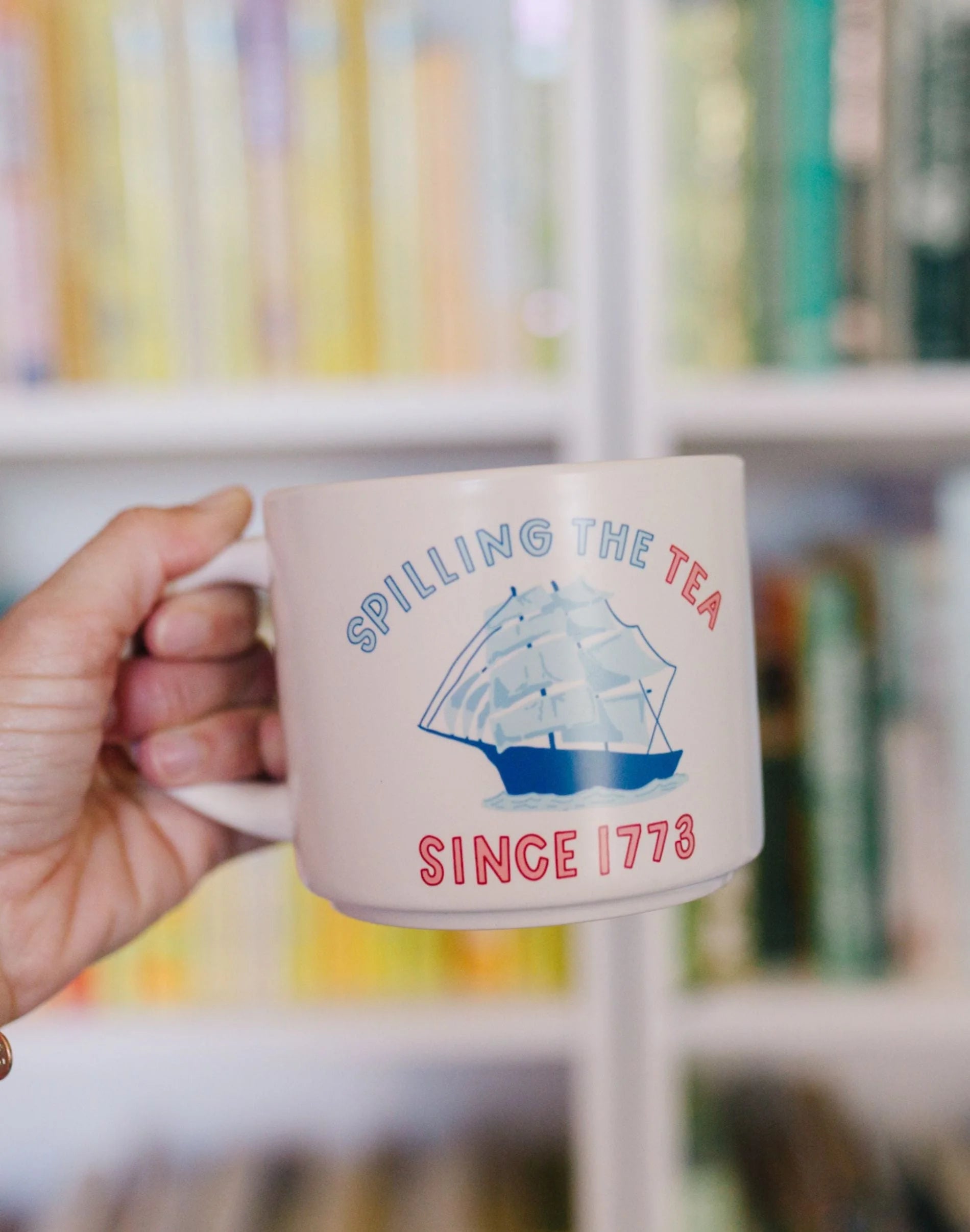 Mug with 'Spilling the Tea Since 1773' design held by a hand in front of a bookshelf.