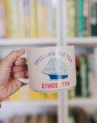 Mug with 'Spilling the Tea Since 1773' design held by a hand in front of a bookshelf.