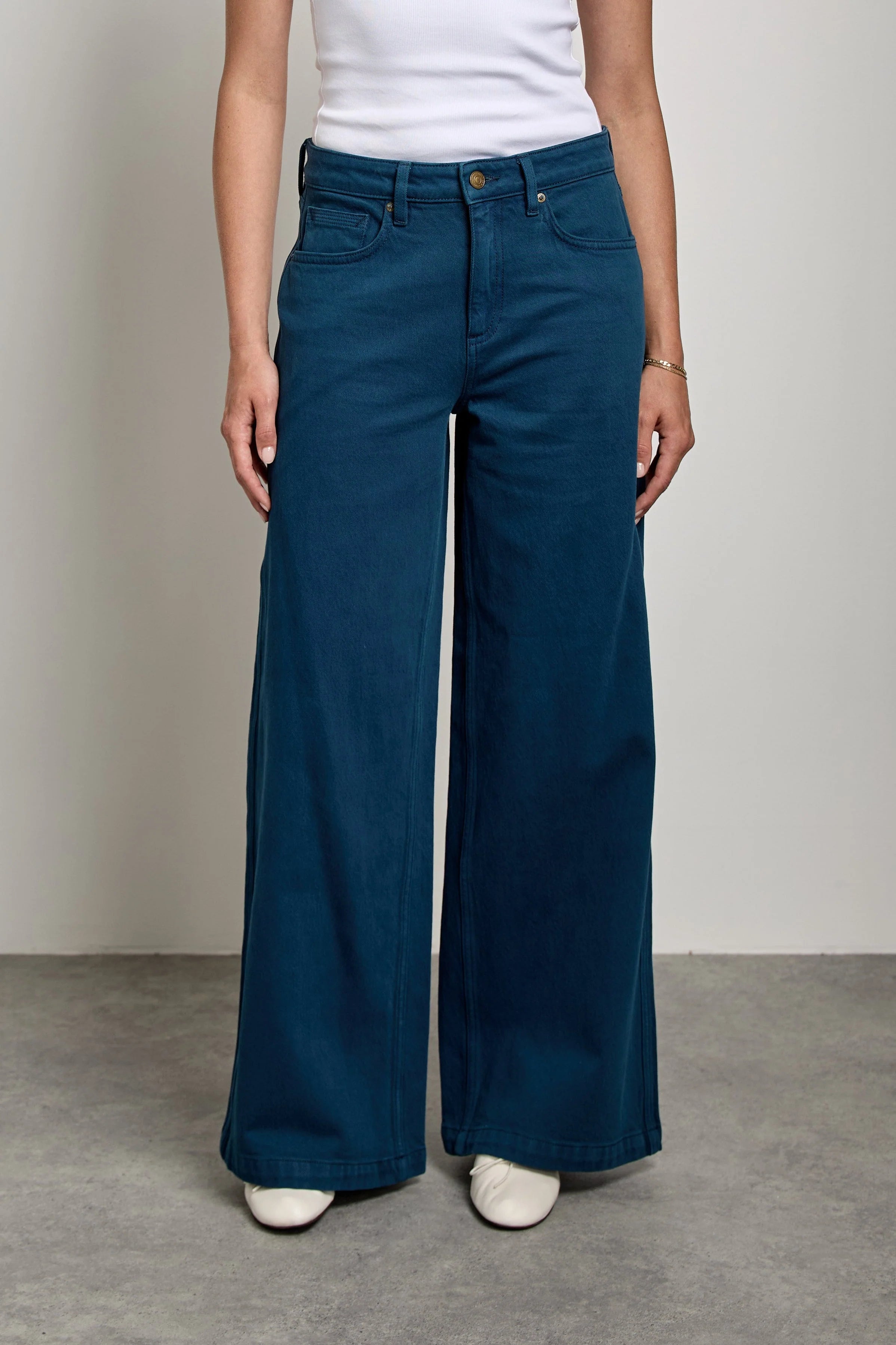 A woman stands in a studio wearing a white ribbed tank top and dark teal wide-leg pants with white sneakers, photographed against a light gray wall and concrete floor.