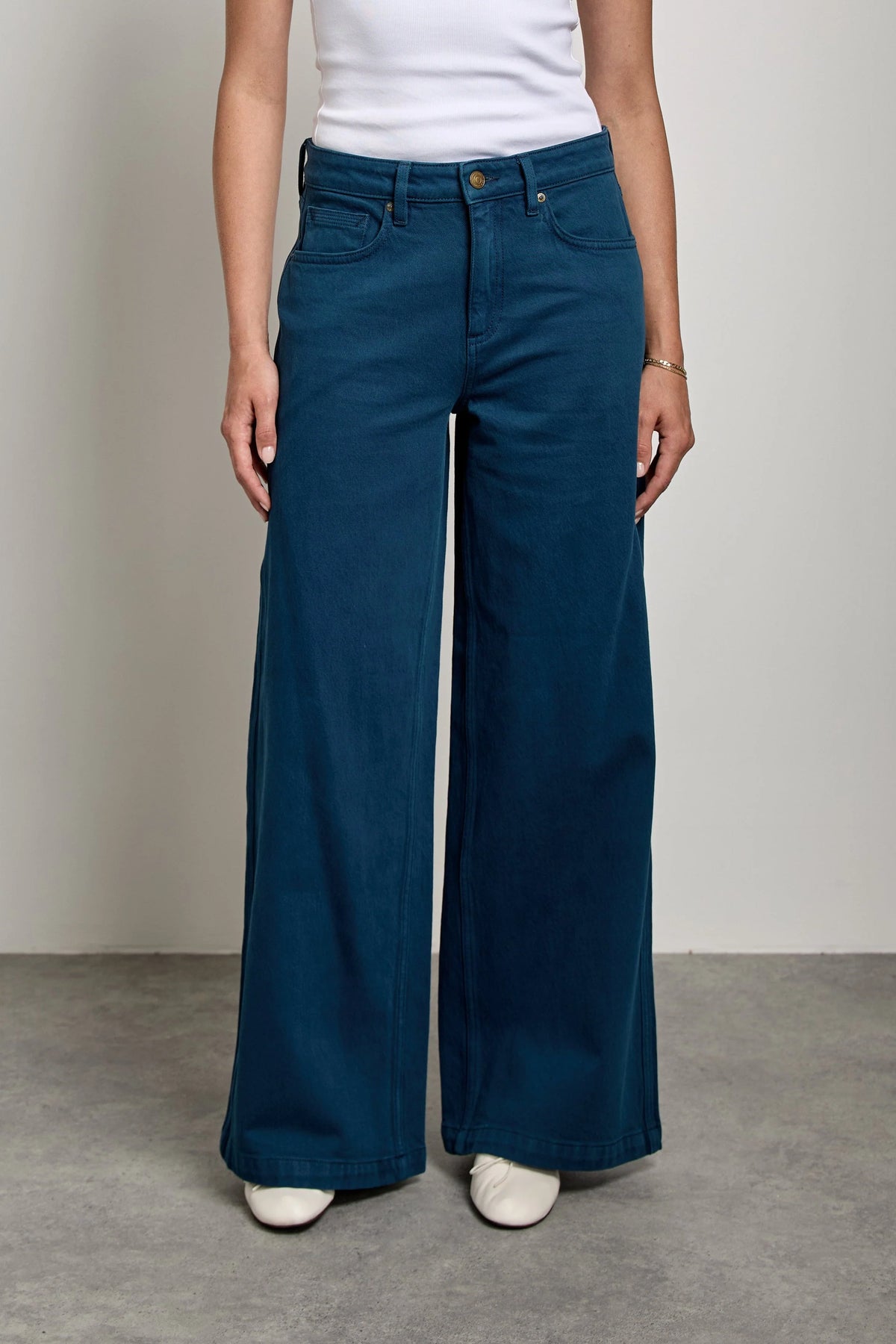 A woman stands in a studio wearing a white ribbed tank top and dark teal wide-leg pants with white sneakers, photographed against a light gray wall and concrete floor.