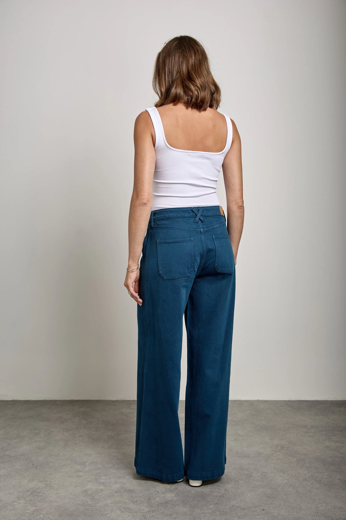 A woman shown from the back wearing wide-leg blue trousers and a white tank top, standing against a neutral studio backdrop.