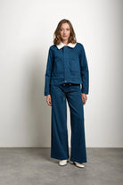 A woman stands in a studio wearing a dark blue denim jacket with a contrasting white collar and matching wide-leg denim pants, paired with white shoes.