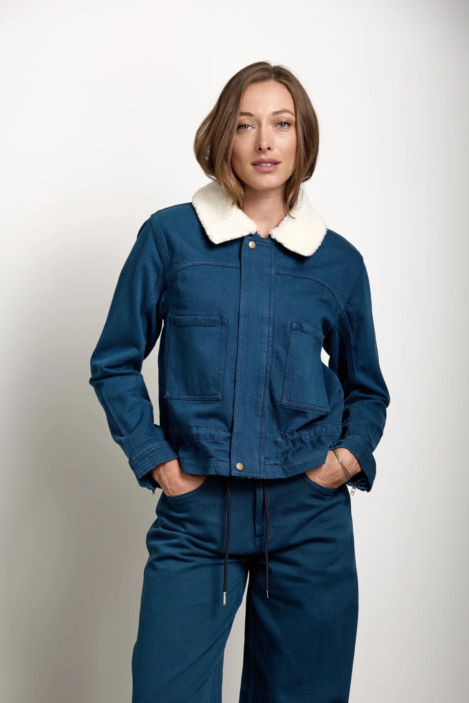 A woman modeling a dark blue denim jacket with a cream-colored shearling-style collar, paired with matching dark blue denim pants, against a plain light gray studio background.