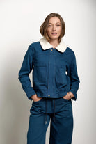A woman modeling a dark blue denim jacket with a cream-colored shearling-style collar, paired with matching dark blue denim pants, against a plain light gray studio background.