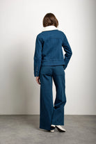 A model viewed from behind wearing a dark blue denim jacket and matching wide-leg pants with a white collar lining, standing on a gray concrete floor against a white wall.