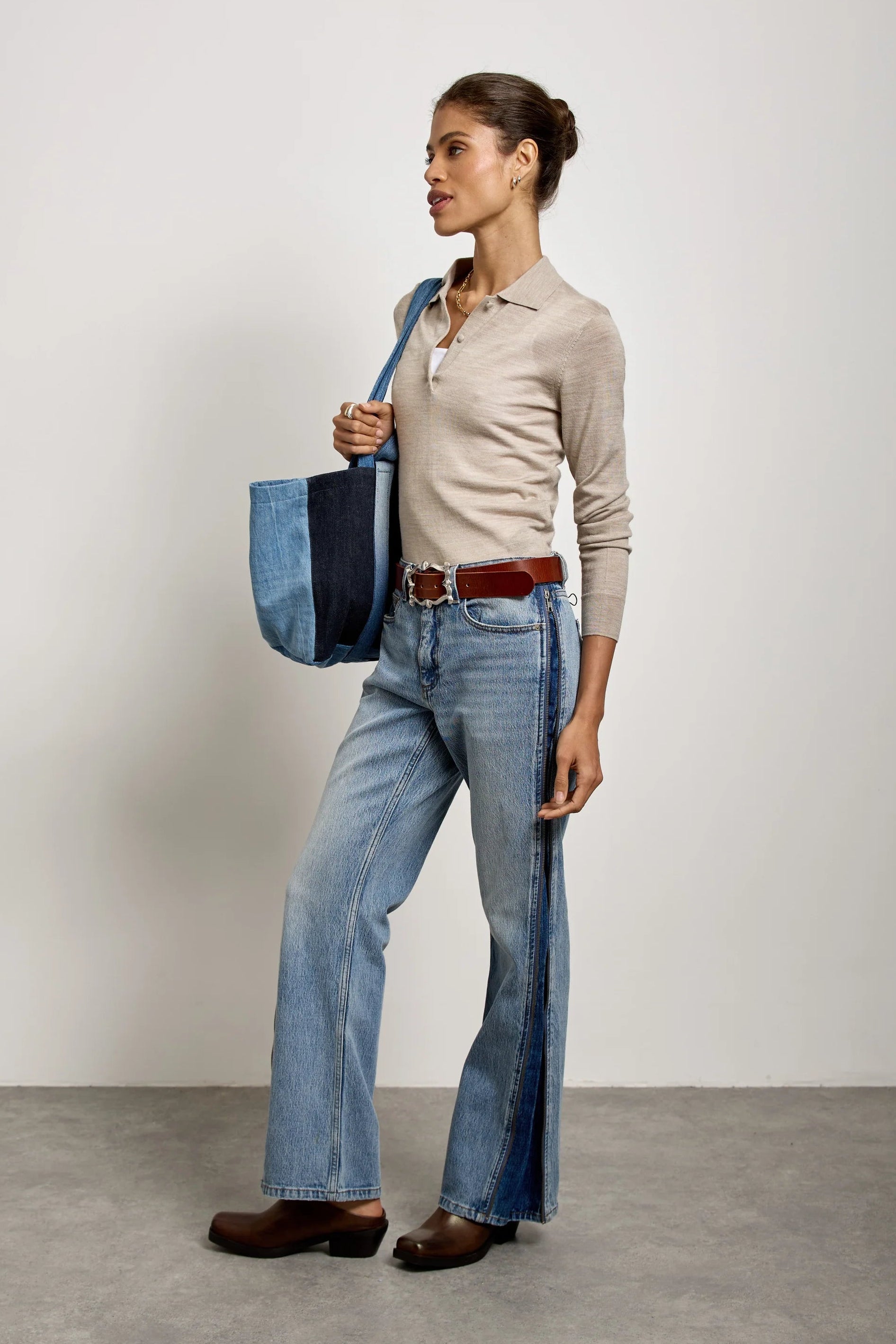 A woman stands in profile wearing light blue wide-leg jeans with dark side panels, a beige long-sleeve polo shirt, a brown belt, and brown shoes, while holding a blue tote bag against a plain light gray studio background.