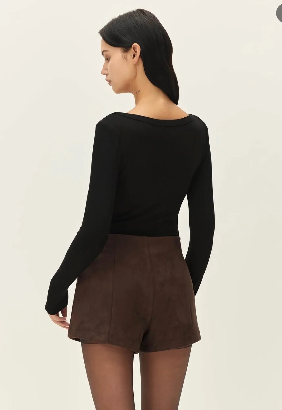 A woman shown from the back wearing a fitted black long-sleeve top and brown shorts against a plain light background.