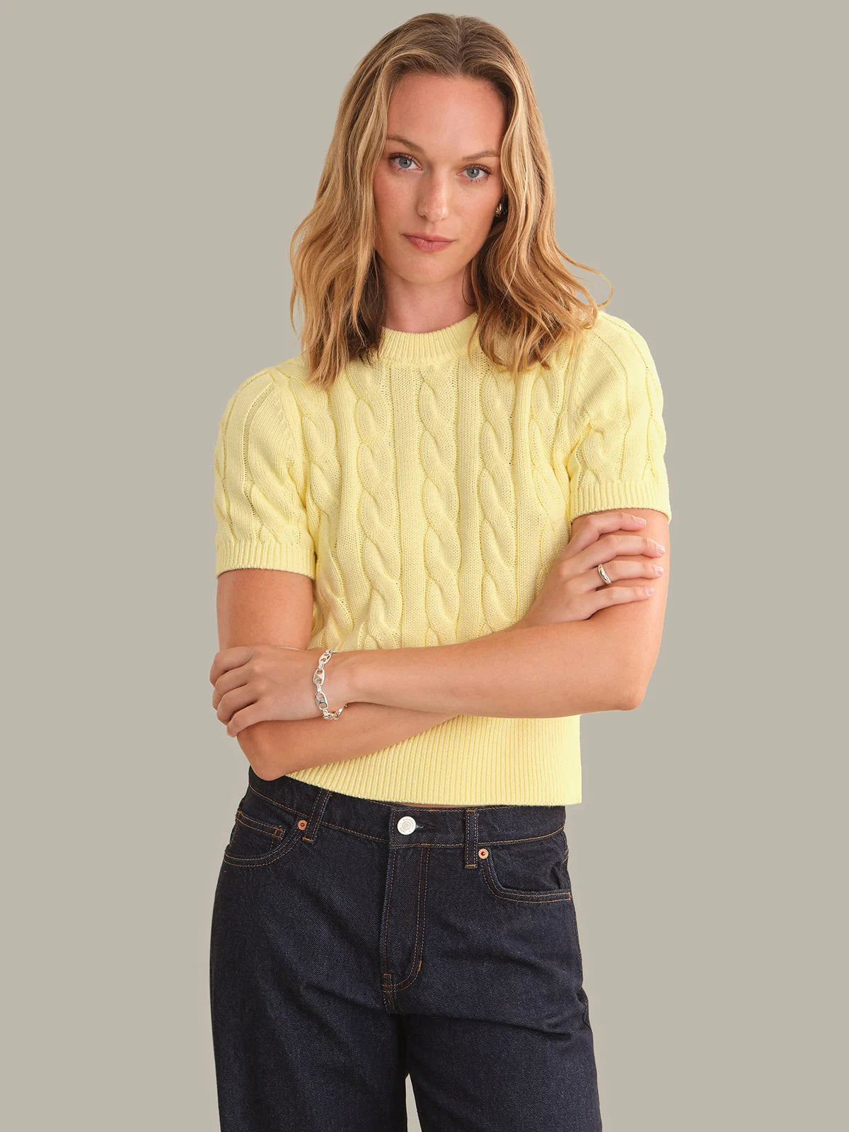 A woman modeling a pale yellow short-sleeve cable-knit sweater with dark blue jeans, standing against a solid gray studio background.