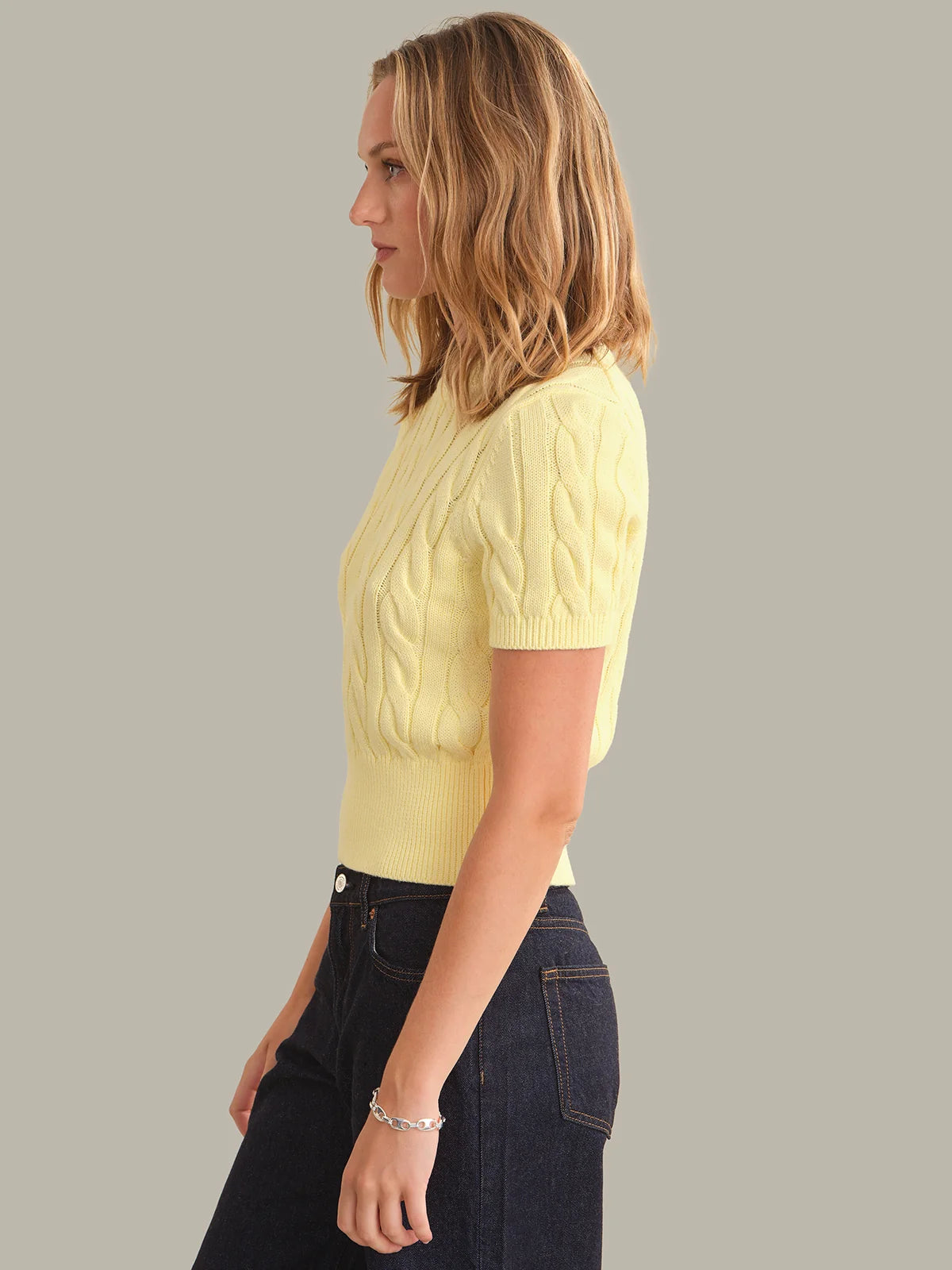 A woman shown in side profile wearing a pale yellow short-sleeve cable-knit sweater and dark blue jeans against a solid light gray studio background.