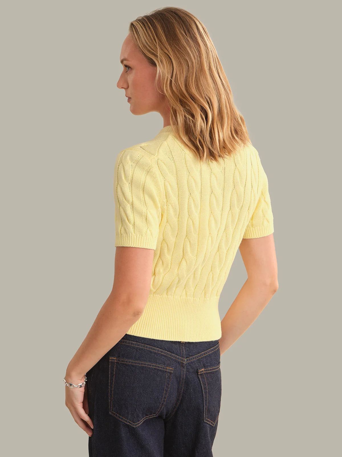 A woman shown from the back wearing a pale yellow short-sleeve cable-knit sweater and dark blue jeans, set against a solid light gray studio background.