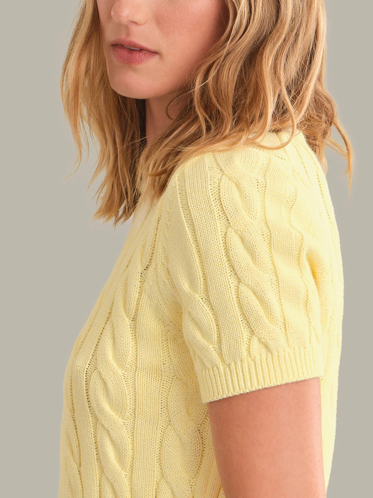 A close-up side view of a woman wearing a pale yellow cable-knit short-sleeve sweater against a plain light gray background.