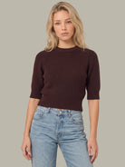 A woman modeling a dark brown ribbed knit sweater with three-quarter sleeves and light-wash blue jeans, set against a solid neutral gray background.
