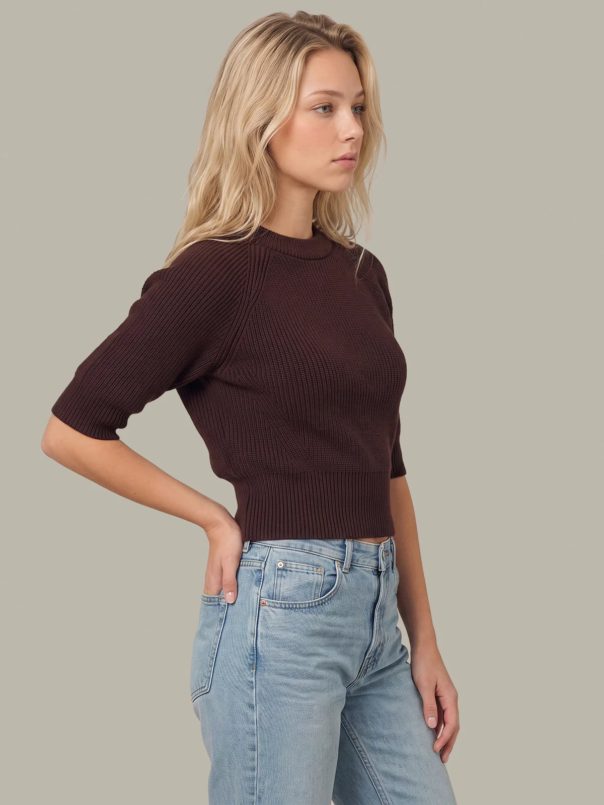 A woman wearing a dark brown ribbed short-sleeve crop top and light blue jeans, standing against a solid light gray background.