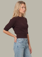 A woman wearing a dark brown ribbed short-sleeve crop top and light blue jeans, standing against a solid light gray background.