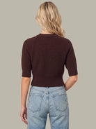 A woman shown from the back wearing a dark brown ribbed knit short-sleeve top and light blue jeans, set against a solid neutral gray background.