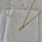 A gold chain necklace with a small rectangular gold tag pendant and a tiny clear stone, laid on a textured off-white linen cloth over a light stone surface.