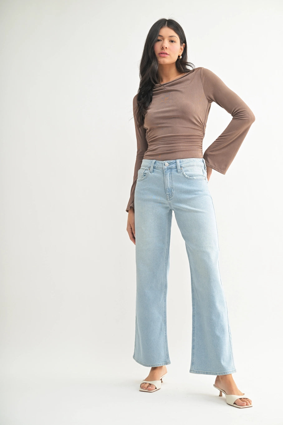 A woman modeling a brown long-sleeve draped top and light-wash wide-leg jeans against a plain light gray studio background.