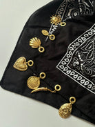A black bandana scarf with a white paisley print, styled with multiple gold-toned metal charms including a sun, shell, heart, coin, crescent moon, and teardrop pendant.