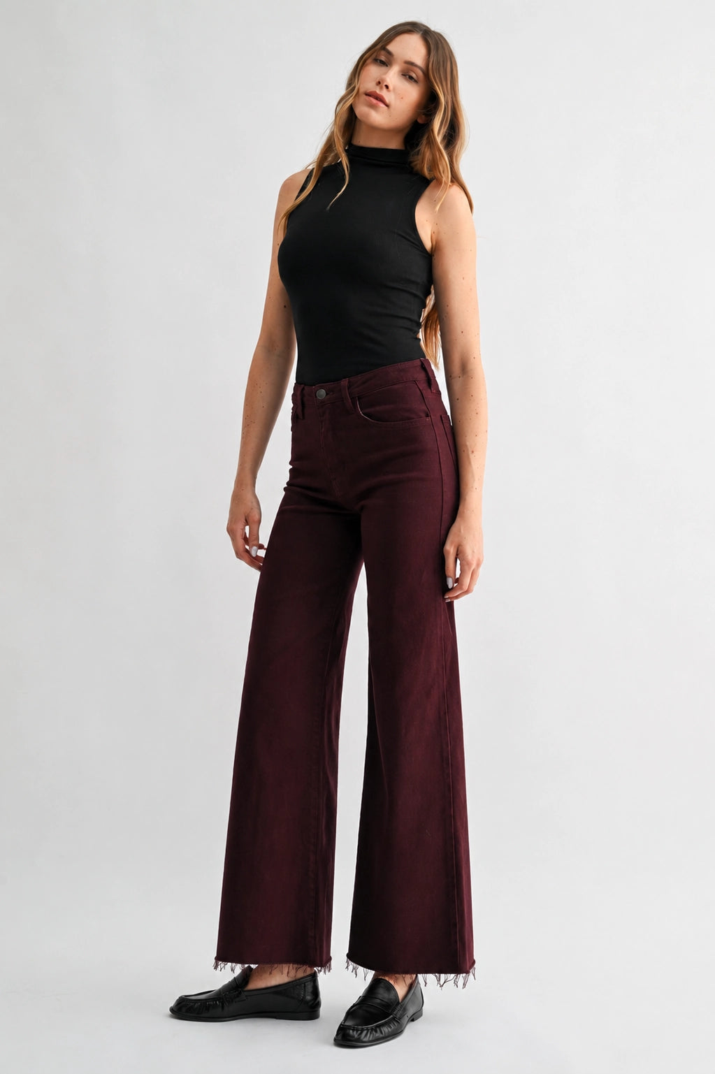A woman modeling a pair of high-waisted, wide-leg burgundy pants with a frayed hem, styled with a black sleeveless top and black loafers against a light gray studio background.