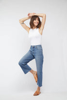Woman wearing a white sleeveless top and blue jeans standing against a plain background