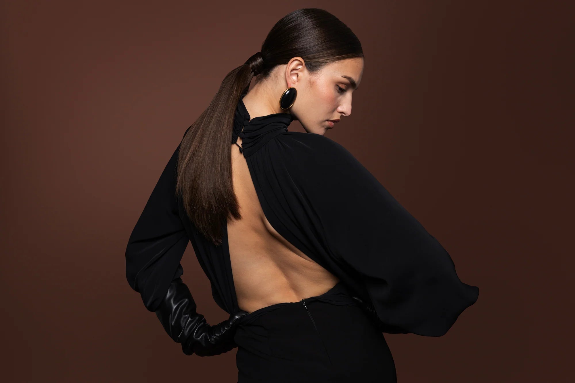 A woman with her hair in a ponytail is shown from the back wearing a black open-back dress with dramatic draped sleeves and black gloves, posed against a solid dark brown studio background.