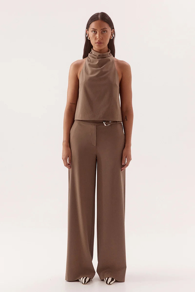 A model stands against a plain light background wearing a matching taupe-brown sleeveless mock-neck top and wide-leg trousers set with a thin belt.