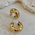 Gold crescent-shaped hoop earrings with heart-shaped cutouts, displayed on a textured light beige stone surface.