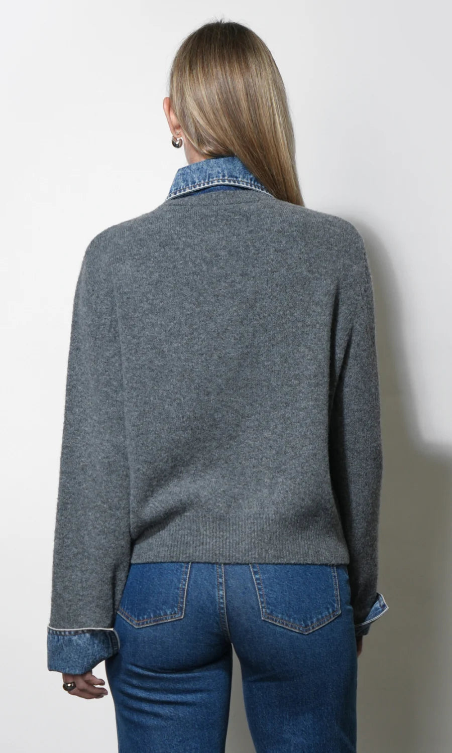 A woman shown from the back wearing a heather gray knit sweater layered over a blue denim collared shirt and blue jeans, against a plain off-white wall.